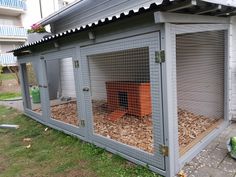 a small grey chicken coop on the side of a building with wood chips in it