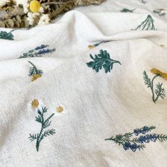 a white blanket with blue and yellow flowers on it, next to dried plants in the background