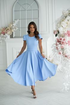 a woman in a blue dress posing for the camera with flowers around her and a white wall behind her