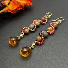 Earthy red creek Jasper rounds in shades of brown, red, and green are wire wrapped in purple, red, and pink seed beads in a swirl pattern in gold wire. Wire wrapped orange Czech glass teardrops dangle from the bottom of the earrings. The perfect fall statement earrings. The earrings measure approximately 3.25 inches from the top of the gold plated ear wires. At checkout, you may choose either an organza gift bag, or a gift box. Fall Earrings Beaded, Earthy Earrings, Red Creek Jasper, Jasper Earrings, Fall Earrings, Earrings Beaded, Jewellery Ideas, Earrings Red, Swirl Pattern