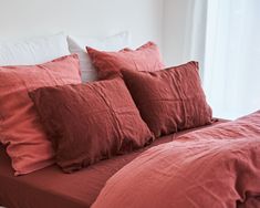 an unmade bed with red linens and white pillows on the headboard, in front of a window