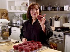 a woman holding up a chocolate cupcake in front of some red velvet muffins