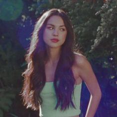 a woman with long dark hair wearing a green top and black pants standing in front of trees