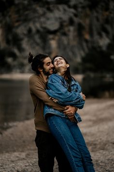 a man and woman embracing each other on the shore of a lake with mountains in the background