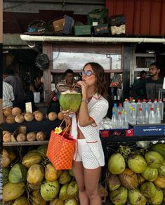 a woman standing next to a pile of coconuts holding a drink in her hand