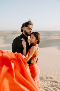 a man and woman standing in the sand with an orange cloth draped around them,