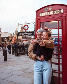 two people standing in front of a red phone booth on the street with their arms around each other