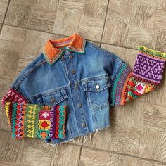 a denim jacket with colorful crocheted patches on the sleeves and cuffs, sitting on a wood floor