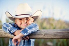 a young boy wearing a cowboy hat leaning on a wooden fence with his arms crossed