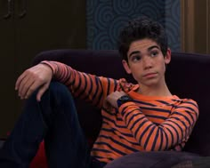 a young man sitting on top of a purple couch next to a black cat and wearing an orange striped shirt