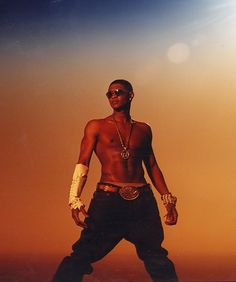 a man with no shirt standing in front of a sky background wearing sunglasses and an arm chain