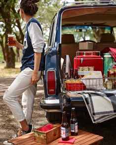 a woman standing next to a car filled with food and drinks while holding a beer