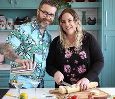 a man and woman standing in front of a cutting board with cheese on it, smiling at the camera