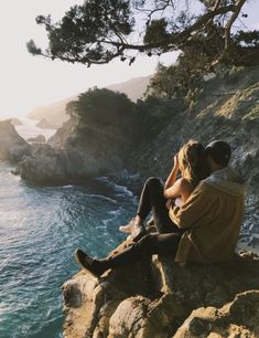 two people sitting on the edge of a cliff looking out at the ocean