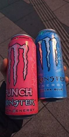 two cans of monster energy drink on the street in front of a person's hand