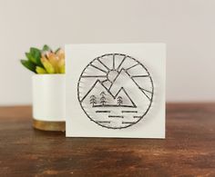 a white card with an image of mountains and trees in the center on a table next to a potted succulent plant