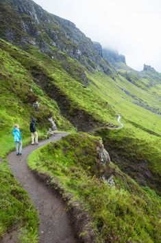 two people walking down a path in the middle of a lush green mountain side area