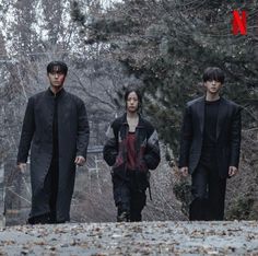 three people are walking down the street in front of some trees and bushes, one is wearing a black coat