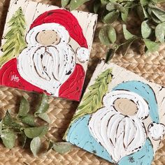 two paintings of santa claus on wood are sitting next to green leaves and plants, one is painted red and the other is white