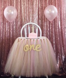 a pink and gold one birthday cake sitting on top of a chair with balloons in the background