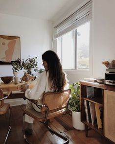 a woman sitting at a table in front of a window with plants on the side