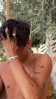 a shirtless man sitting on a wooden bench next to an olive tree with tattoos