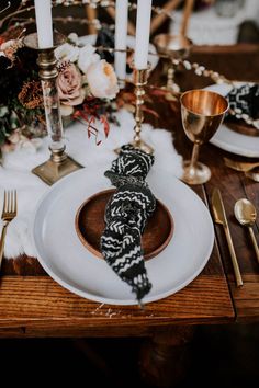 a white plate topped with a black and white scarf next to gold place settings on a wooden table