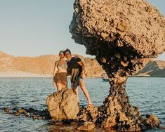 a man and woman standing on top of a rock next to the ocean with mountains in the background