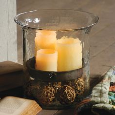 three lit candles in a glass container on top of a table next to an open book