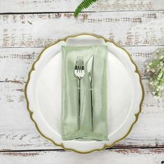 a white plate topped with a green napkin and fork next to a fern plant on top of a wooden table