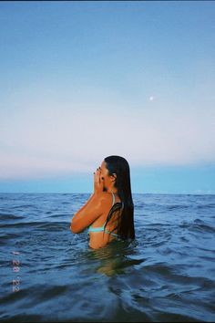 a woman standing in the water with her hand to her face and looking up at the sky