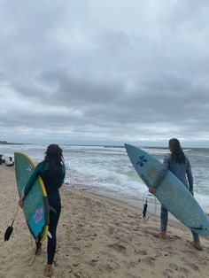 two women carrying surfboards on the beach with waves in the background and people walking by