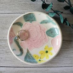 a ring dish with flowers and leaves painted on the side, next to a plant