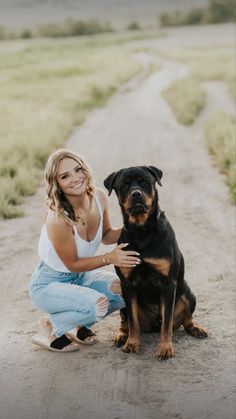 a woman kneeling down next to a black and brown dog on a dirt road with grass in the background