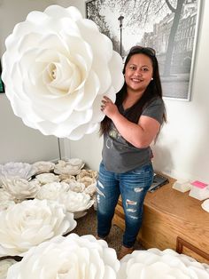 a woman holding a giant white flower in front of a table full of paper flowers