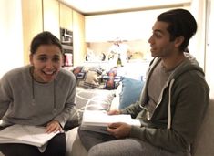 a young man and woman sitting on a couch smiling at each other while holding books