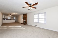 an empty living room with a ceiling fan and brick fireplace in the corner, next to a kitchen