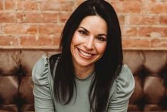 a woman sitting on top of a brown couch next to a brick wall and smiling at the camera