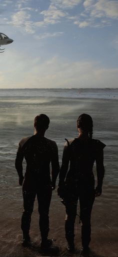 two people standing on the beach looking at an airplane in the sky over the water