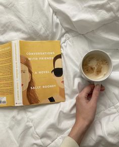 a person is reading a book and holding a cup of coffee while laying on a bed