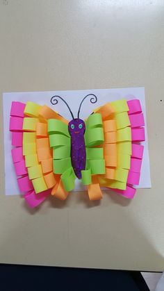 a butterfly made out of paper on top of a piece of white paper with pink, yellow and green strips