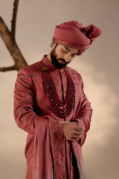 a man with a beard wearing a red suit and beaded necklaces standing in front of a tree