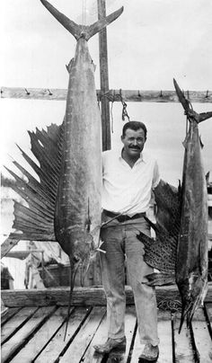 black and white photo of man holding two large fish on dock with fishing poles in background