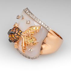 This beautifully fashioned whimsical ring is topped with mother of pearl and accented on the edges with a total of thirty-six (36) bead set round brilliant cut diamonds that are lined with milgrain details. The top of the ring features a black rhodium and rose gold fish, bead set with thirty-six (36) round brilliant cut orange sapphires and eighteen (18) round brilliant cut, light yellow sapphires. The eyes are each bezel set with one (1) round modified brilliant cut tsavorite garnet. The mother Elegant White Multi-stone Diamond Ring, Luxury White Diamond Ring With Accent Stones, Luxury Multi-stone White Rings, Luxury White Jewelry With Accent Stones, Whimsical Ring, Fish Ring, Cut Orange, Tsavorite Garnet, Gold Fish