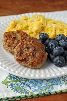 a white plate topped with meat patties and blueberries next to scrambled eggs on a wooden table