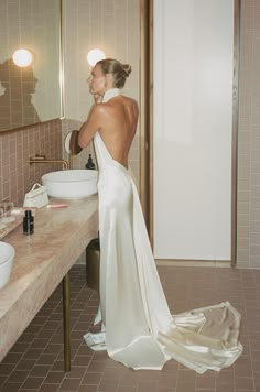 a woman standing in front of a bathroom mirror wearing a white dress and looking at herself in the mirror