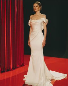 a woman standing in front of a red curtain wearing a white dress with one shoulder