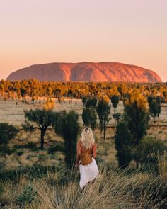 a woman in a white dress walking through tall grass with a mountain in the background