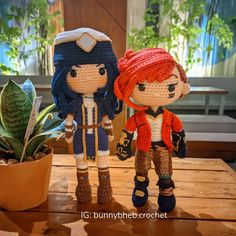 two crocheted dolls standing next to each other on a wooden table in front of a potted plant