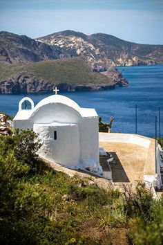 an old white church on the side of a hill with water and hills in the background